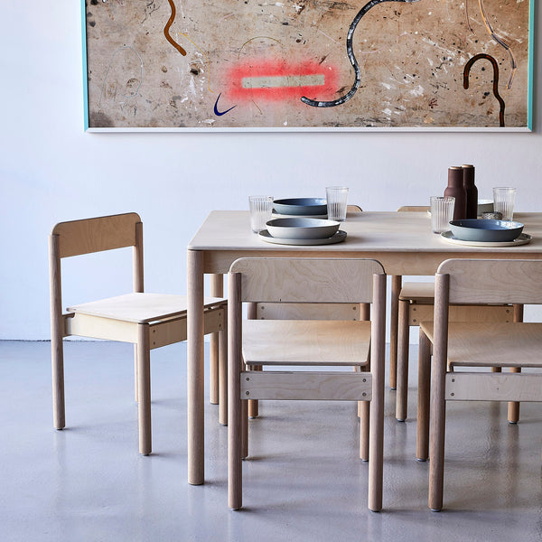 Minimalist Natural Coloured Wooden Dining Table and Chairs with Tablewares on Top and Abstract Painting Behind